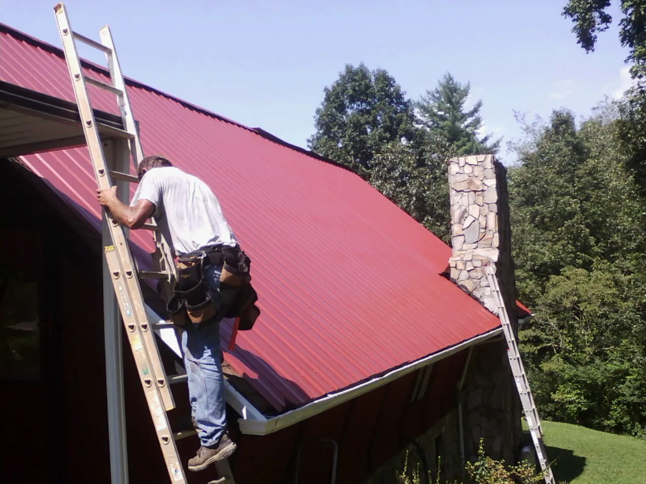 Roofer on a ladder beside a red metal roof and stone chimney — Soffit & Fascia Repair by Roofing Brothers in Seward